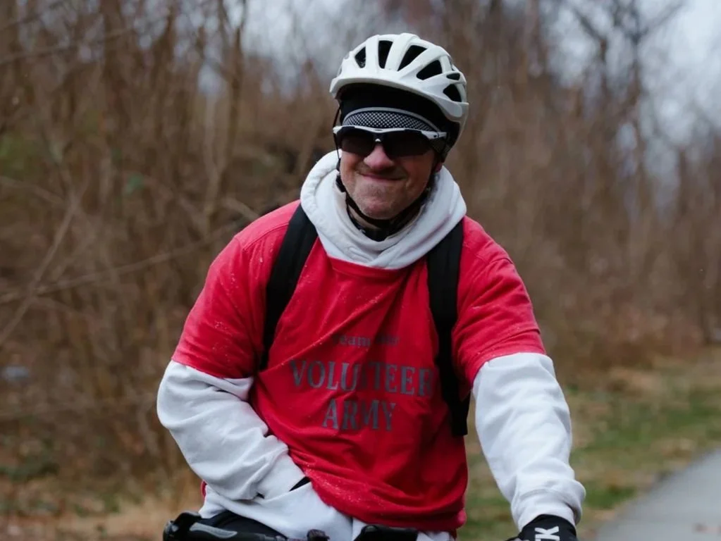 A man wearing a white helmet, sunglasses, and a red sweatshirt that reads 'VOLUNTEER ARMY' with white sleeves, riding a bicycle outdoors with trees in the background.