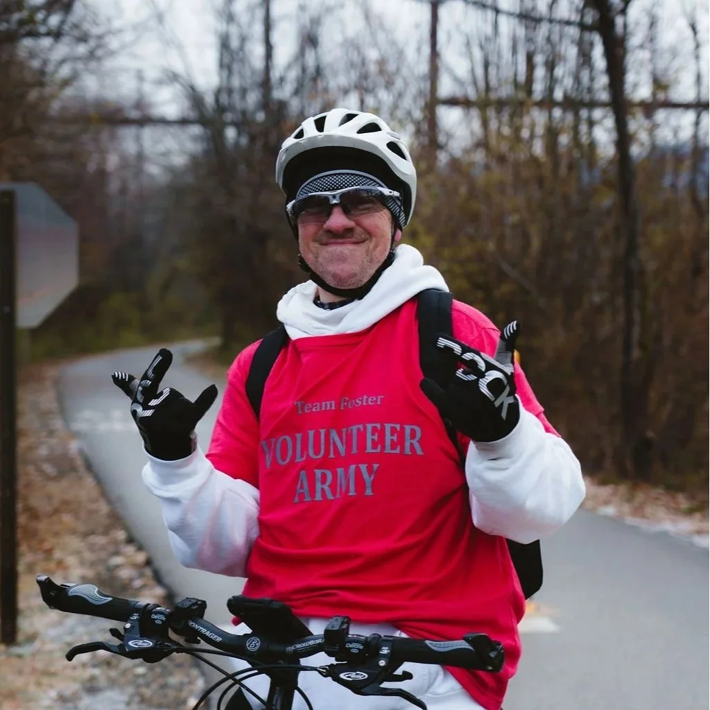 A man wearing a white bicycle helmet, sunglasses, and a red vest with the words 'VOLUNTEER ARMY' on it. He is smiling and making a hand gesture while standing with his bicycle on a trail in a wooded area.