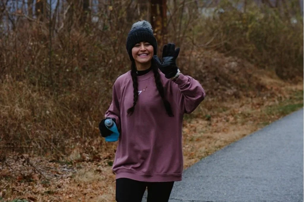Woman in a purple oversized sweatshirt, black beanie, black gloves, braided hair, smiling, walking outdoors on a paved trail in a wooded area, holding a water bottle, waving.