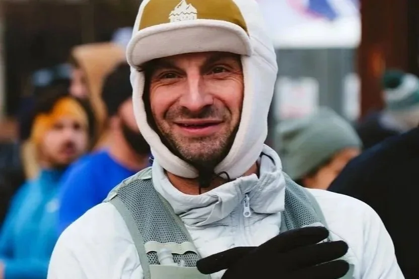 Man smiling in outdoor winter gear, wearing a white hat with ear protection and a light gray jacket, among a crowd.
