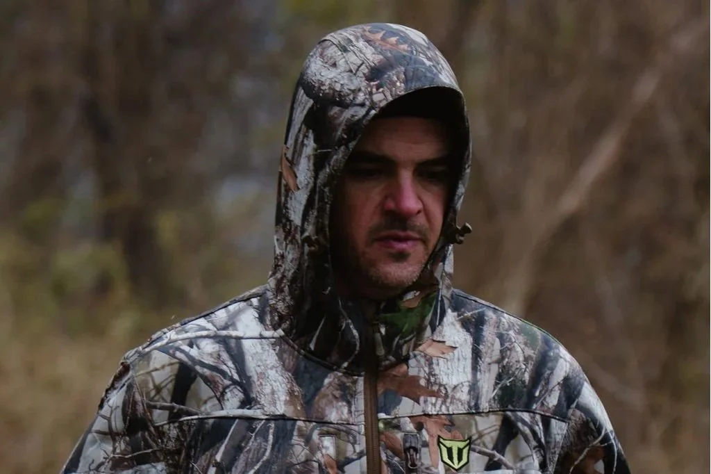 A man wearing camouflage hunting jacket and hood outdoors with trees in the background.