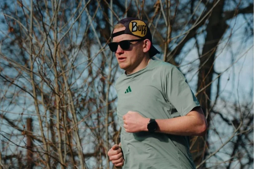 A man running outdoors in a park during daytime, wearing sunglasses, a gray athletic shirt, a black wristwatch, and a black cap with yellow and purple lettering, standing in front of a background of leafless trees.