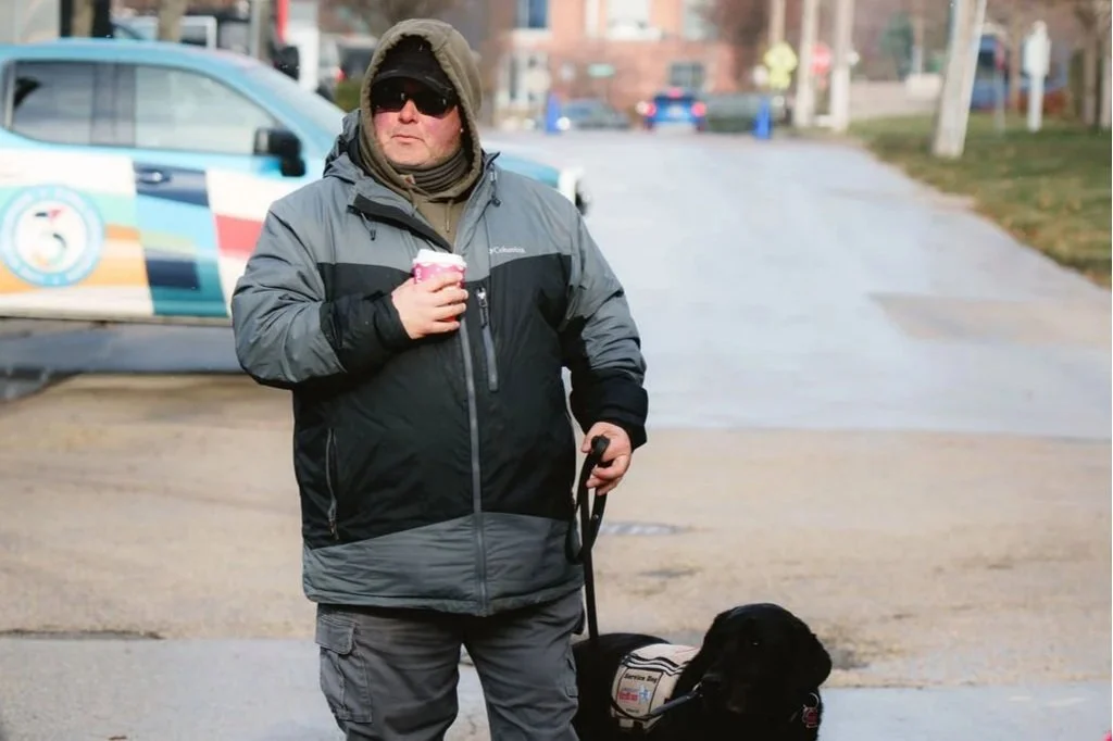 A man in a gray and black jacket, sunglasses, and a hoodie walking a black Labrador Retriever on a leash, holding a coffee cup, on a city sidewalk with a police car in the background.