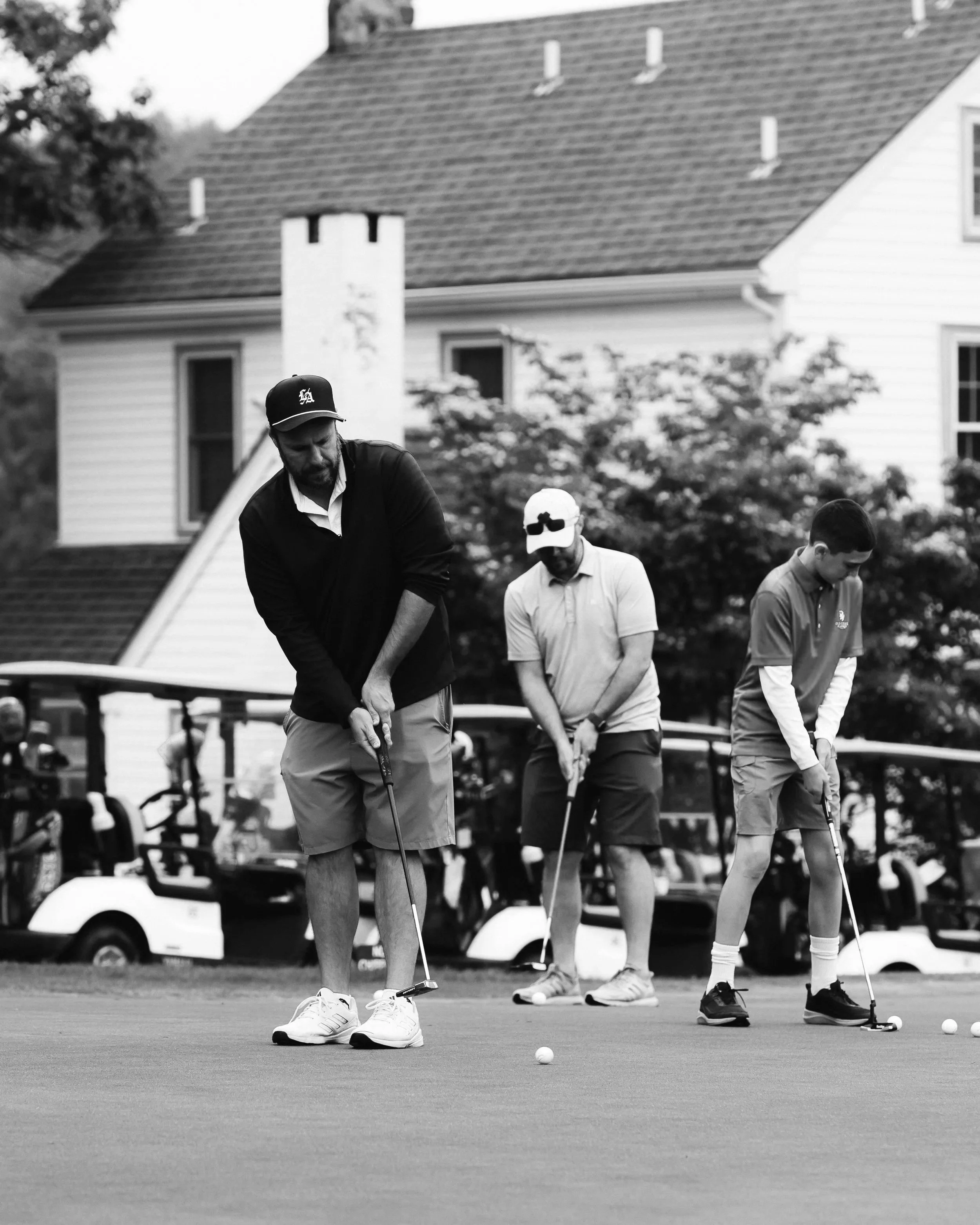 Four men playing golf on a putting green, with a house and golf carts in the background.