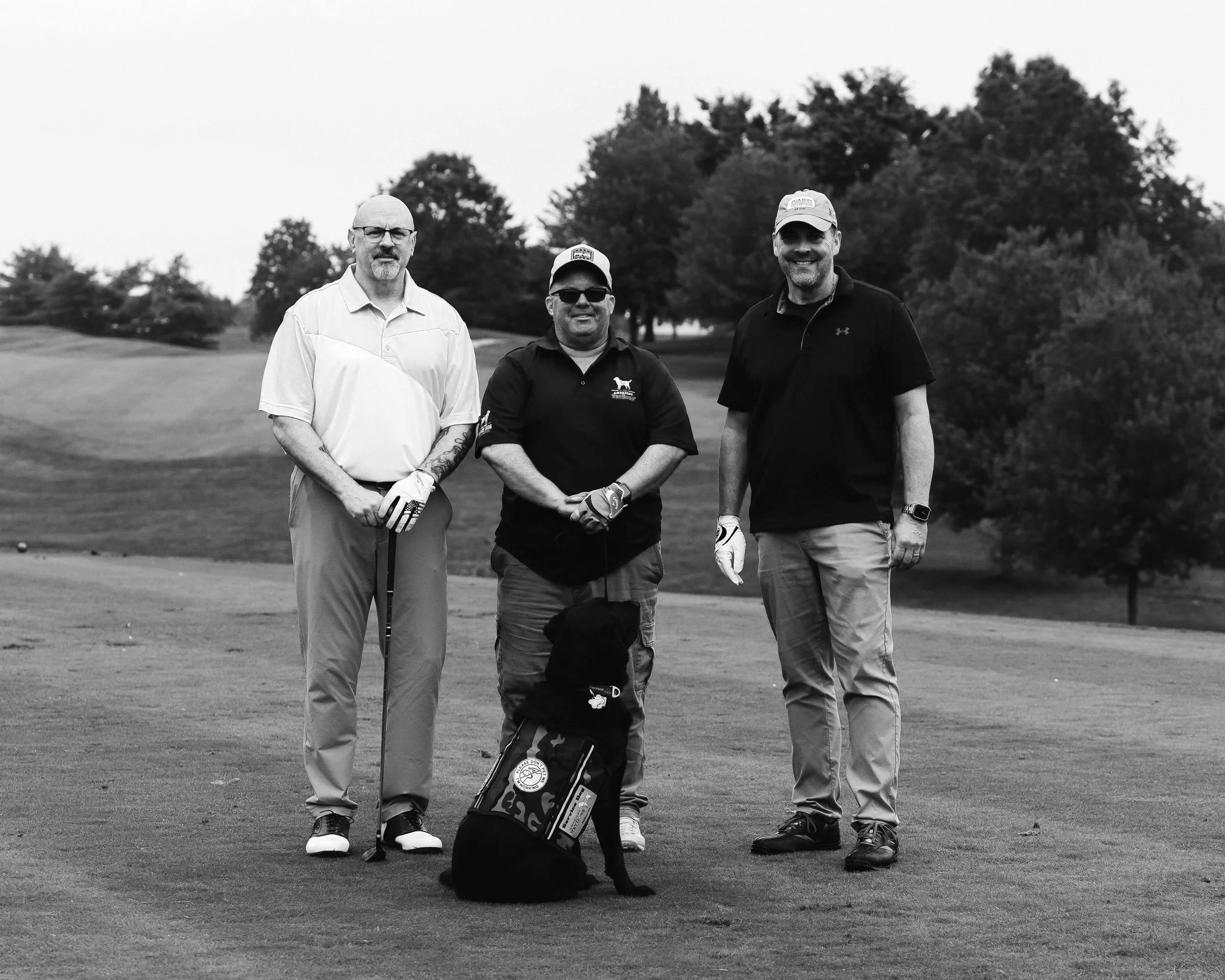 Three men standing on a golf course, smiling, with a dog sitting in front of them. The men are wearing casual golf attire and the background features trees and fairway.