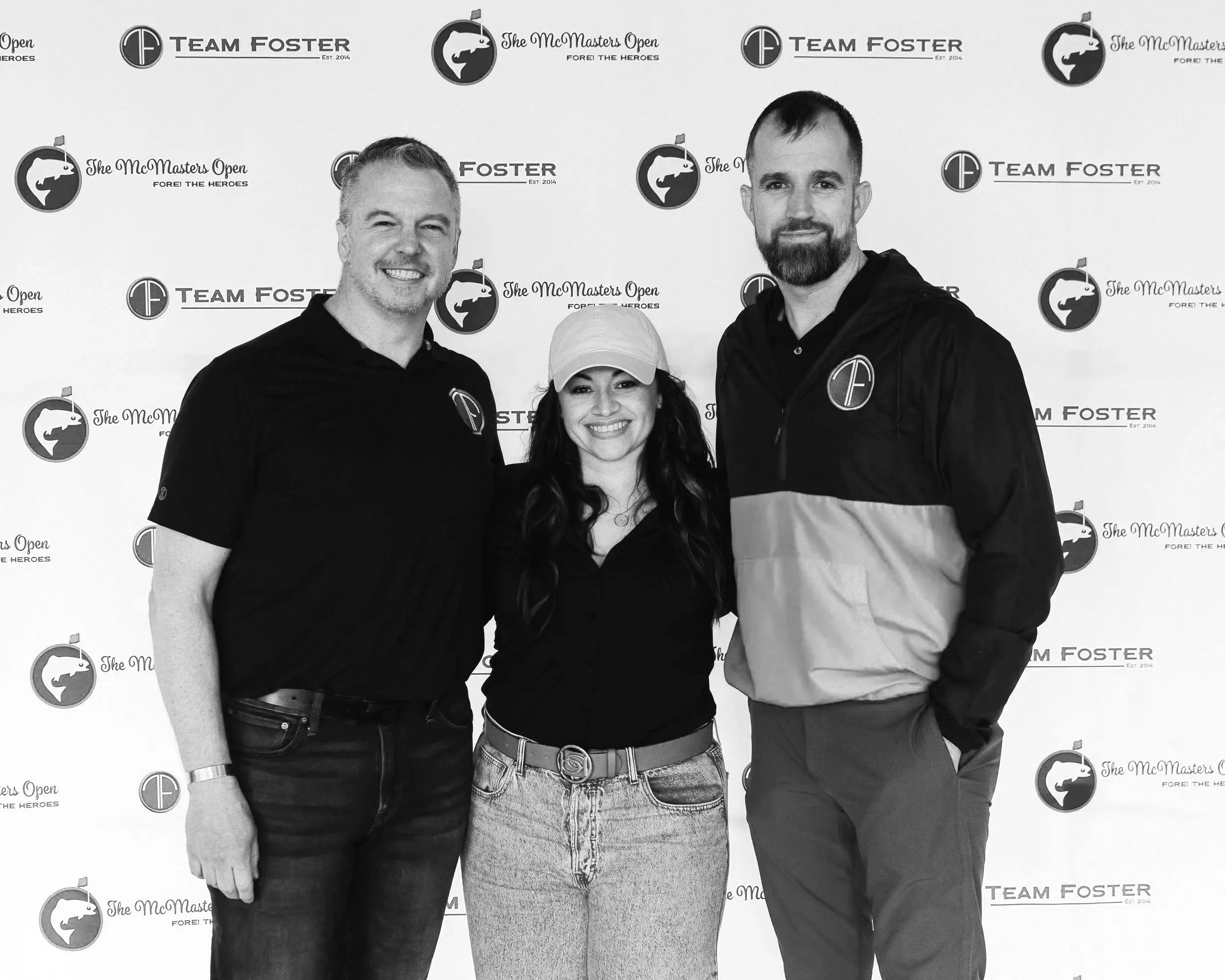 Three people standing in front of a backdrop with logos and text related to 'The McMasters Open' and 'Team Foster'. The group includes two men and one woman, all smiling and dressed casually, with the woman wearing a baseball cap.