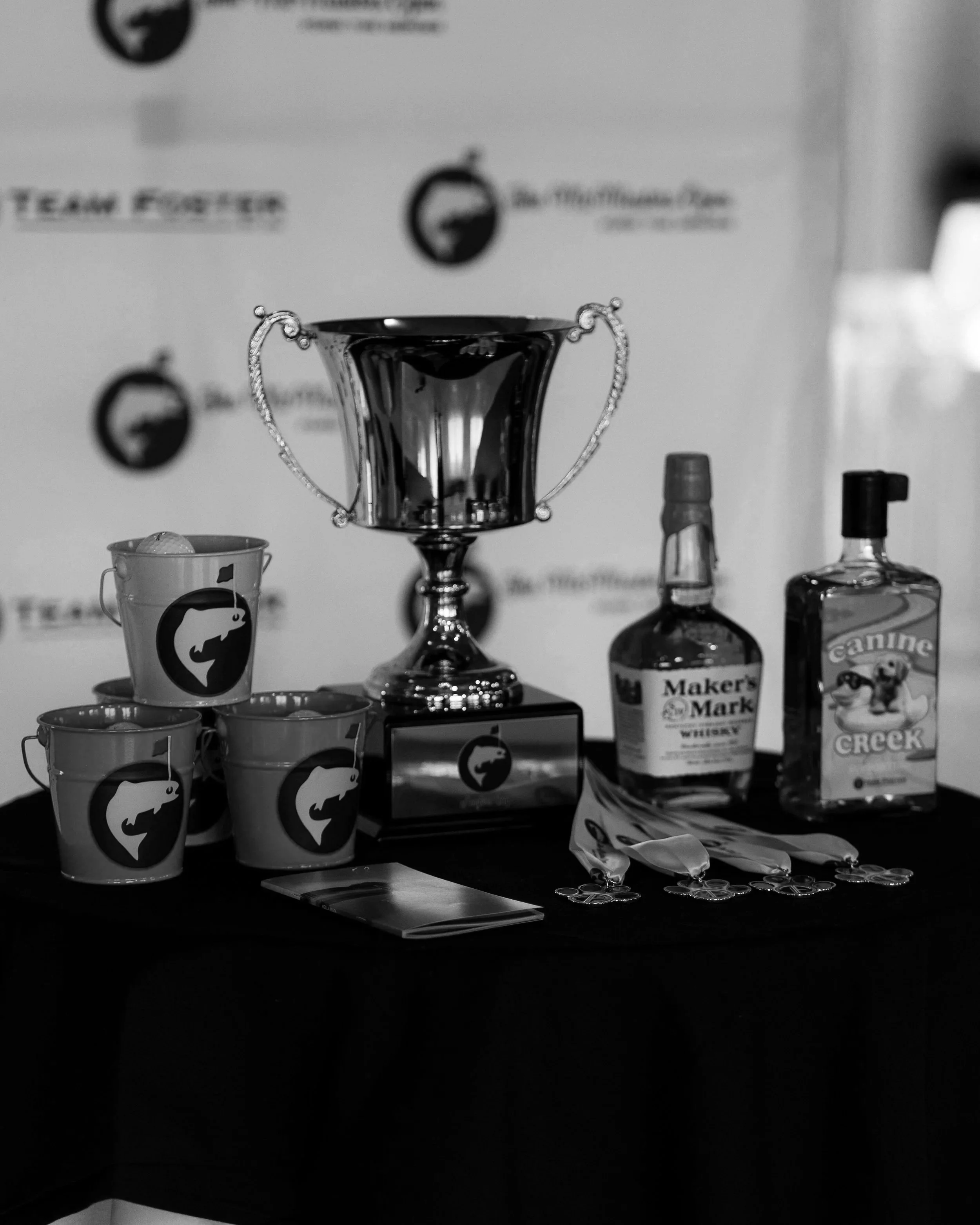 Black and white photo of a display table with several objects, including cups with logos, bottles, a trophy, and various items, with a blurred background.