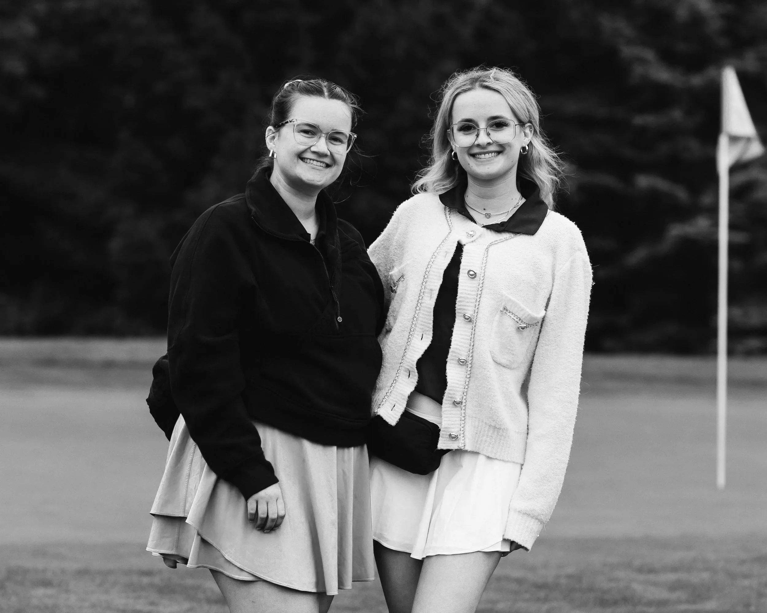 Two young women with glasses and earrings, smiling outdoors, wearing jackets and skirts, standing close together near a flagpole, blurred background of trees.