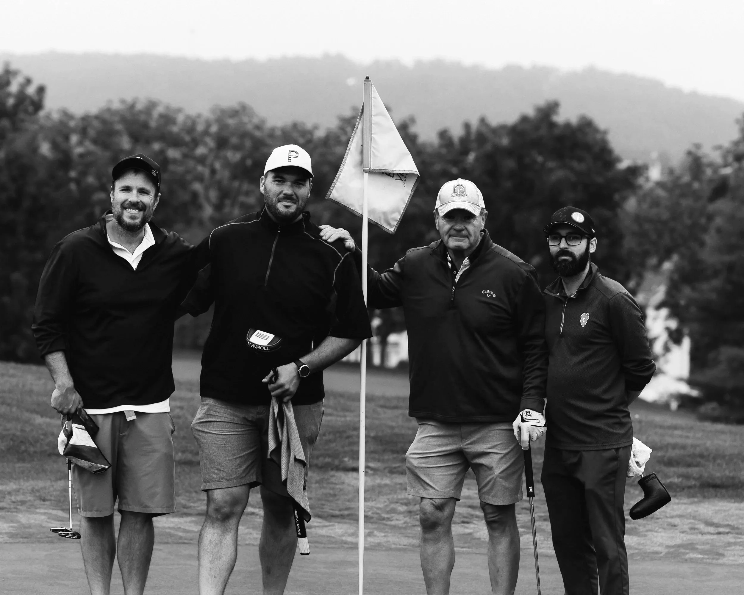 A group of five men standing together on a golf course, with trees and hills in the background. They are dressed in golf attire, and one man is holding a golf flag.