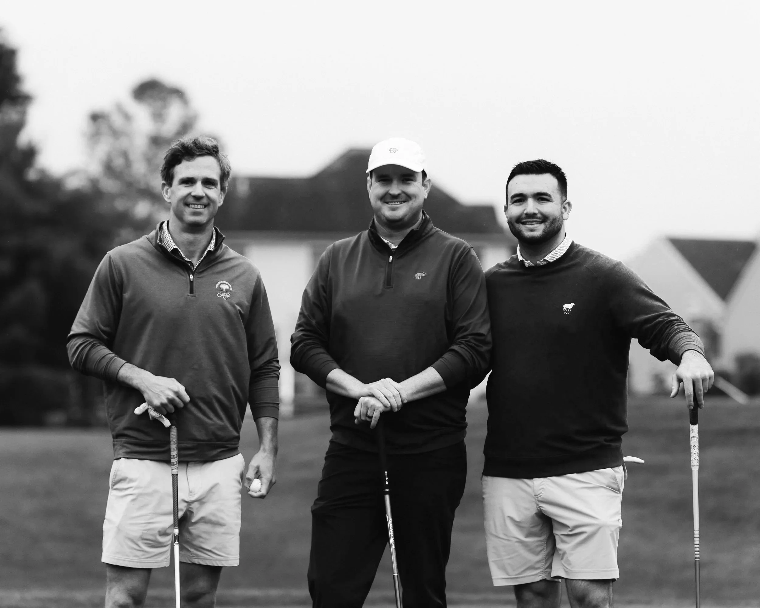 Three men standing on a golf course with golf clubs, smiling, in black and white.