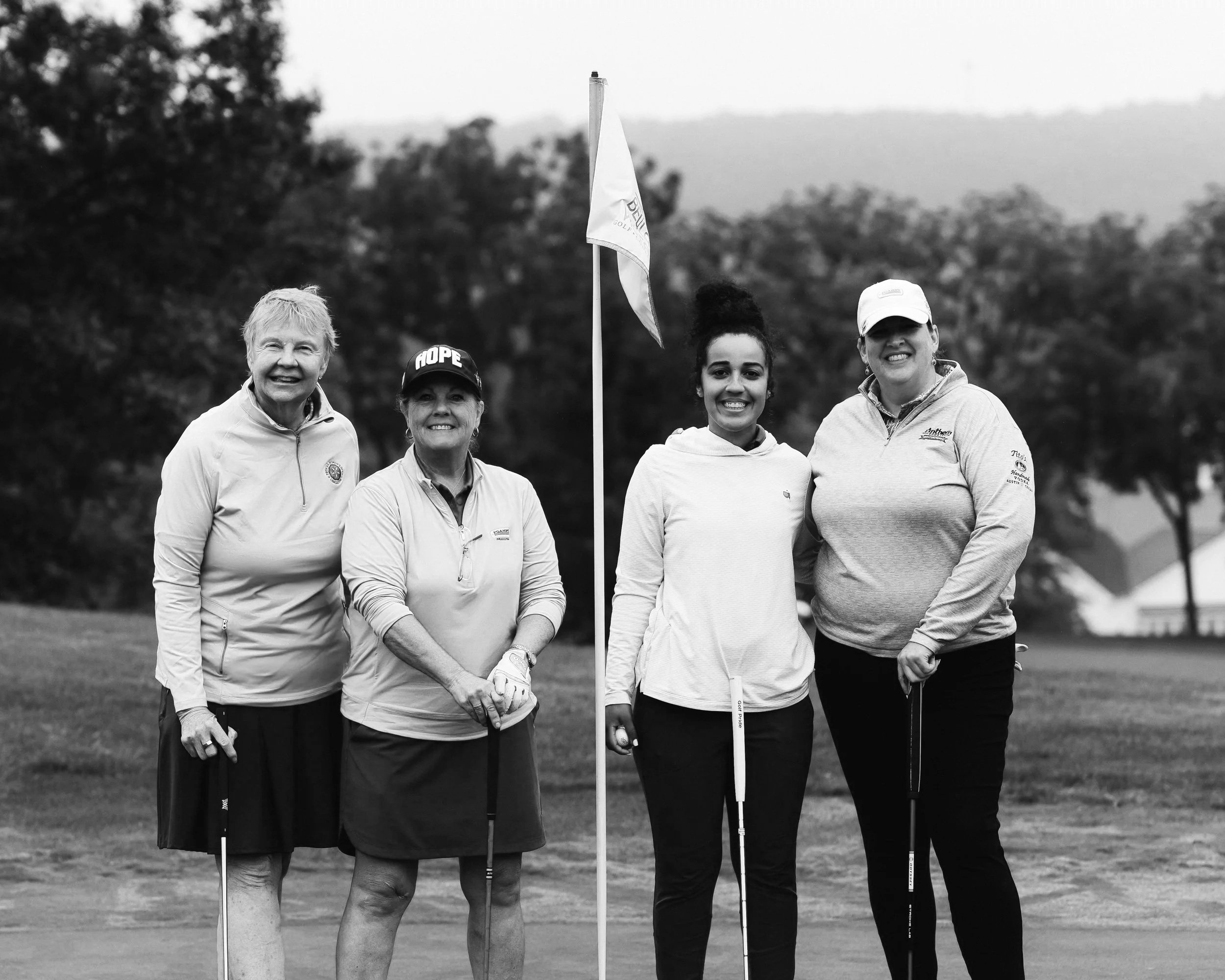 Four women standing on a golf course, holding golf clubs, with a golf flag in the background. They are smiling and wearing athletic clothing.