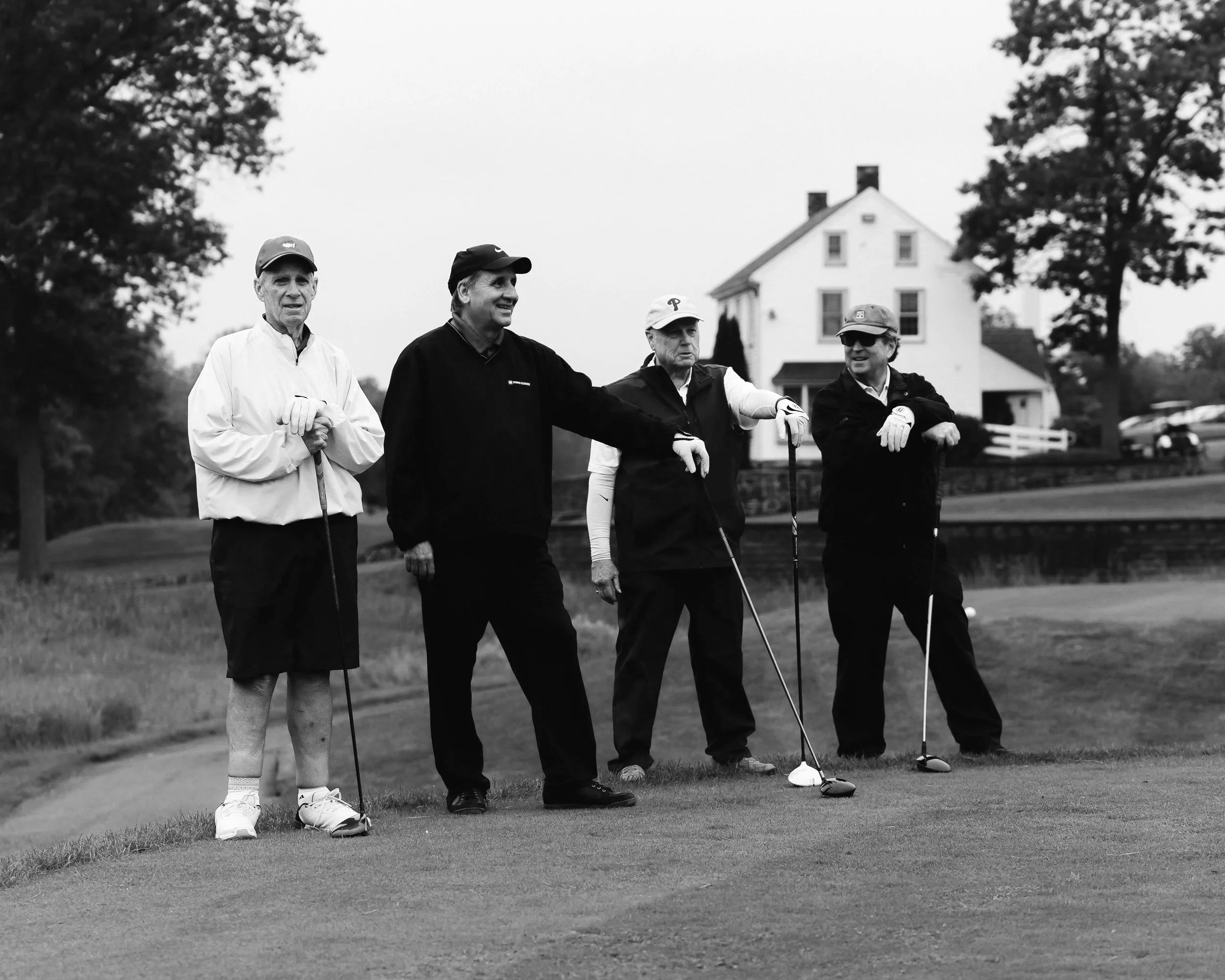 Four men standing on a golf course near a hole with clubs in hand, with a house and trees in the background, in black and white.