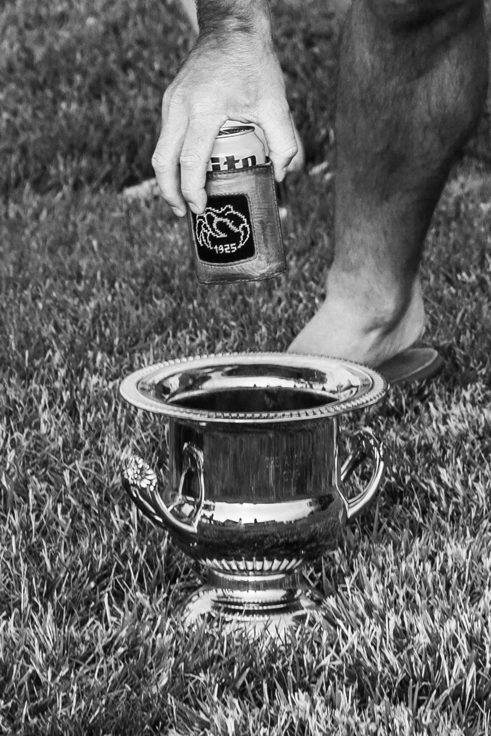 A person is placing a can into a shiny trophy on grass, with part of their leg and foot visible in the background.