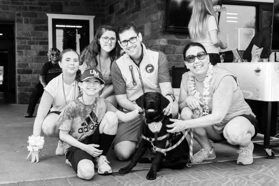 Group of six people and a service dog posing together indoors. The group includes children and adults, with some wearing leis and holding the dog. They are smiling and appear happy.