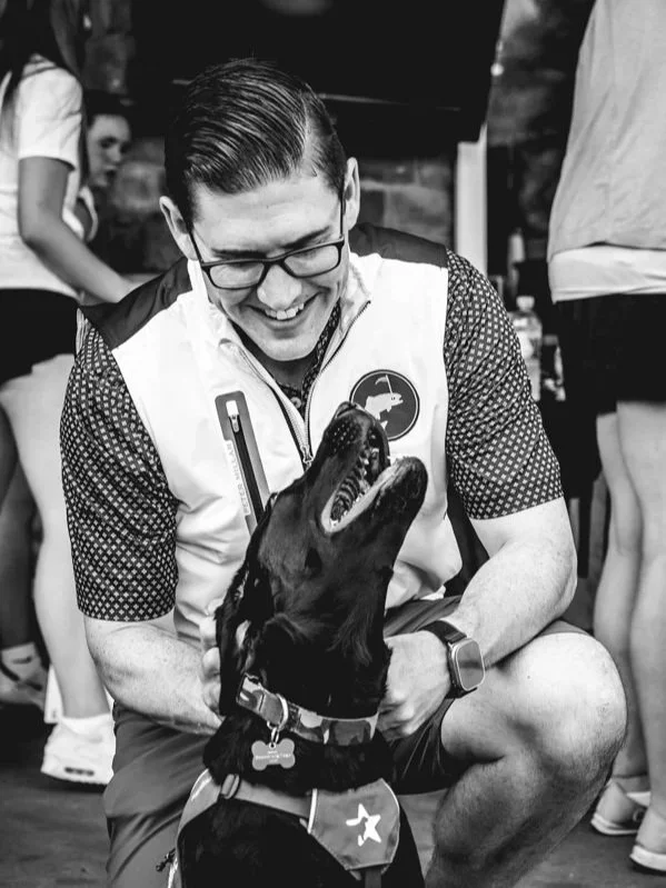 A man wearing glasses and a vest with a patch, smiling while sitting on the ground, engaging with a black service dog with a bone-shaped tag and a vest, at an indoor event with other people around.