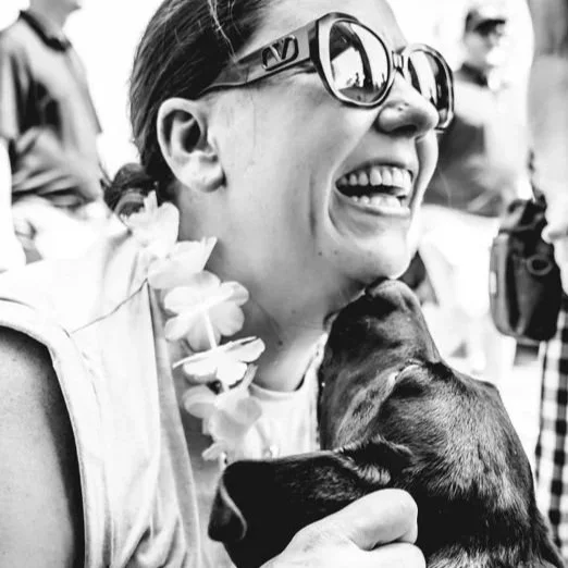 A woman with sunglasses and a floral lei laughing while holding a dog close to her face in black and white.
