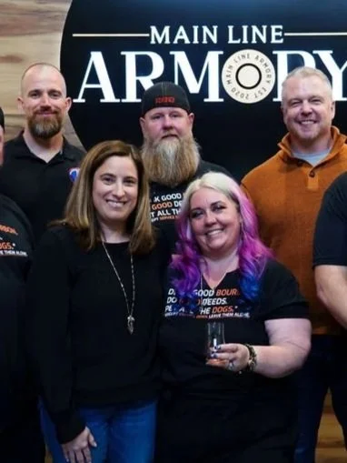Group of five people posing in front of a sign that says 'Main Line Armory', two men and three women, one woman holding a drink, all smiling.