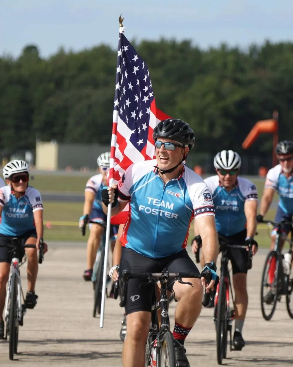 A man rides a bicycle while holding an American flag, part of a group of cyclists wearing