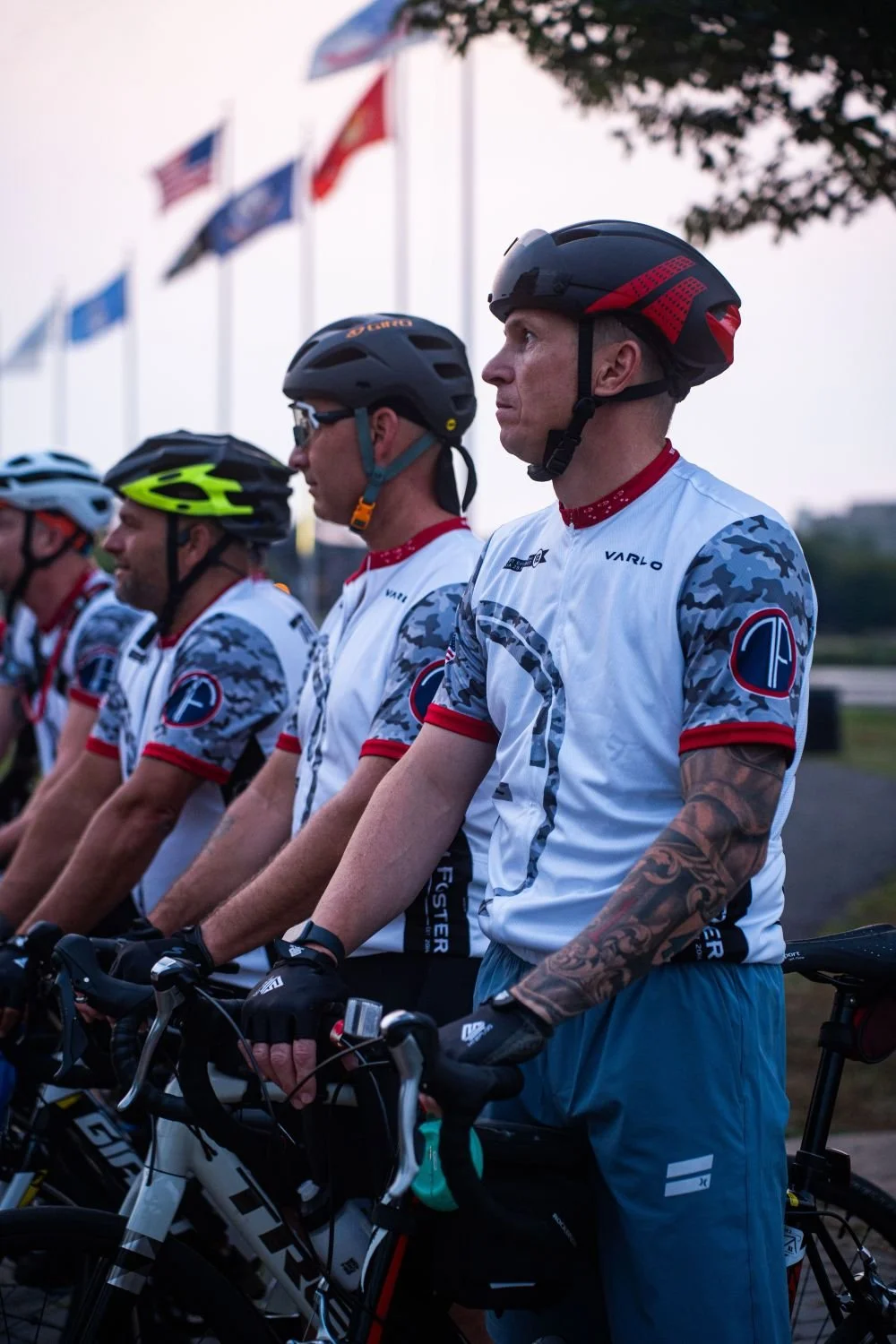 A group of cyclists standing side by side in their cycling gear and helmets, lined up with flags on poles flying in the background during dusk.