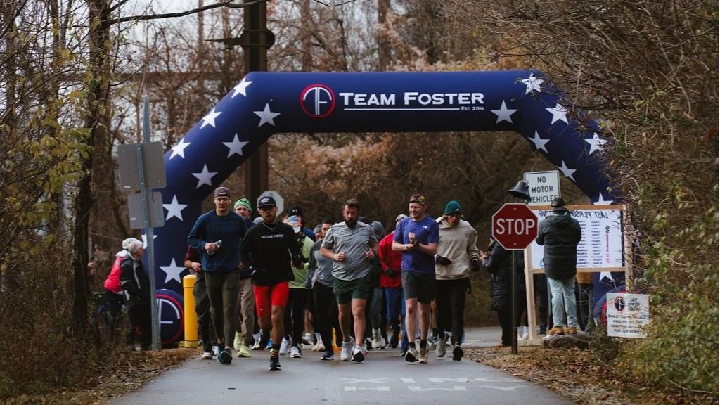 Group of runners participating in a race under a patriotic archway with stars, marked 'Team Foster'. The archway is over a road with a stop sign, a person addressing the runners, and autumn trees lining the background.