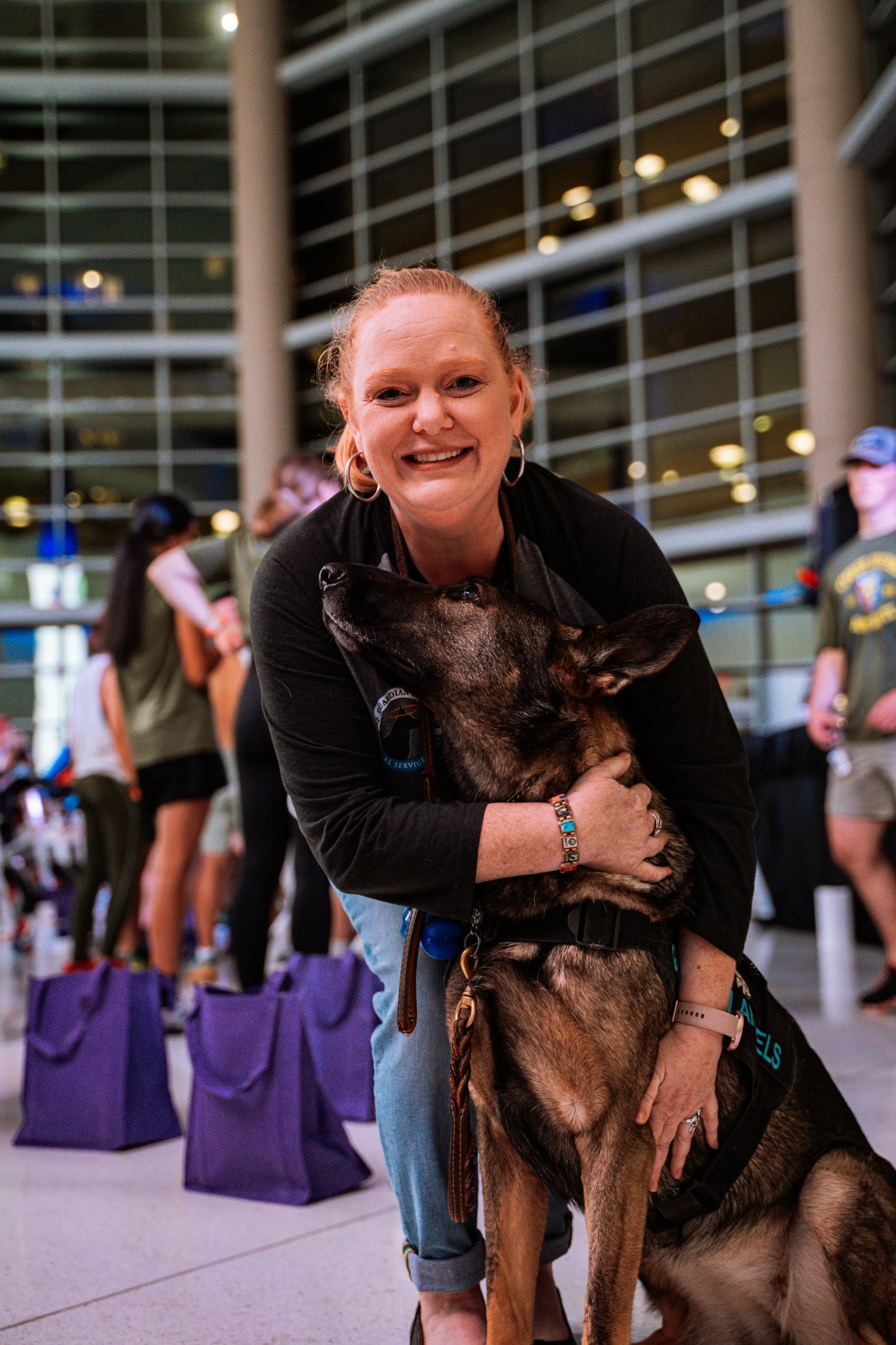 A smiling woman with red hair and hoop earrings hugging a large Belgian Malinois dog at an indoor event. Purple tote bags line the floor behind them.