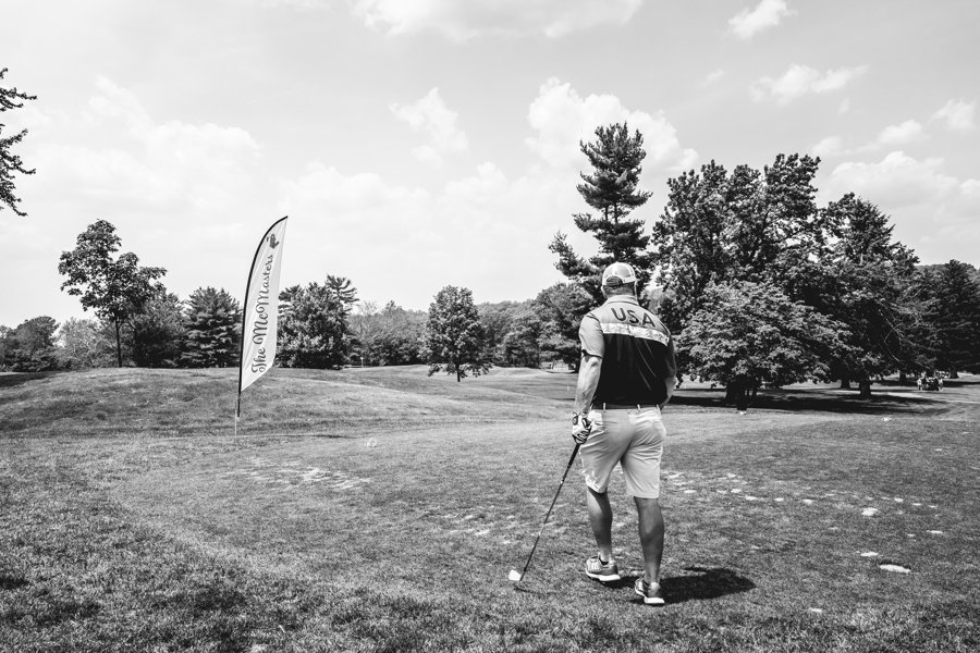 A man in golf attire, wearing a shirt with 'USA' on the back, walking on a golf course with a golf club in his hand, heading towards a flag on the green.