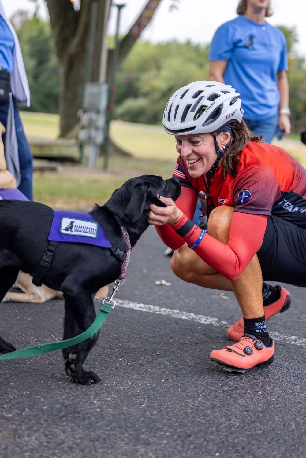 A woman in a red cycling outfit and white helmet crouches on a paved outdoor path, smiling and holding the face of a black service dog wearing a purple vest. There are other people and trees in the background.