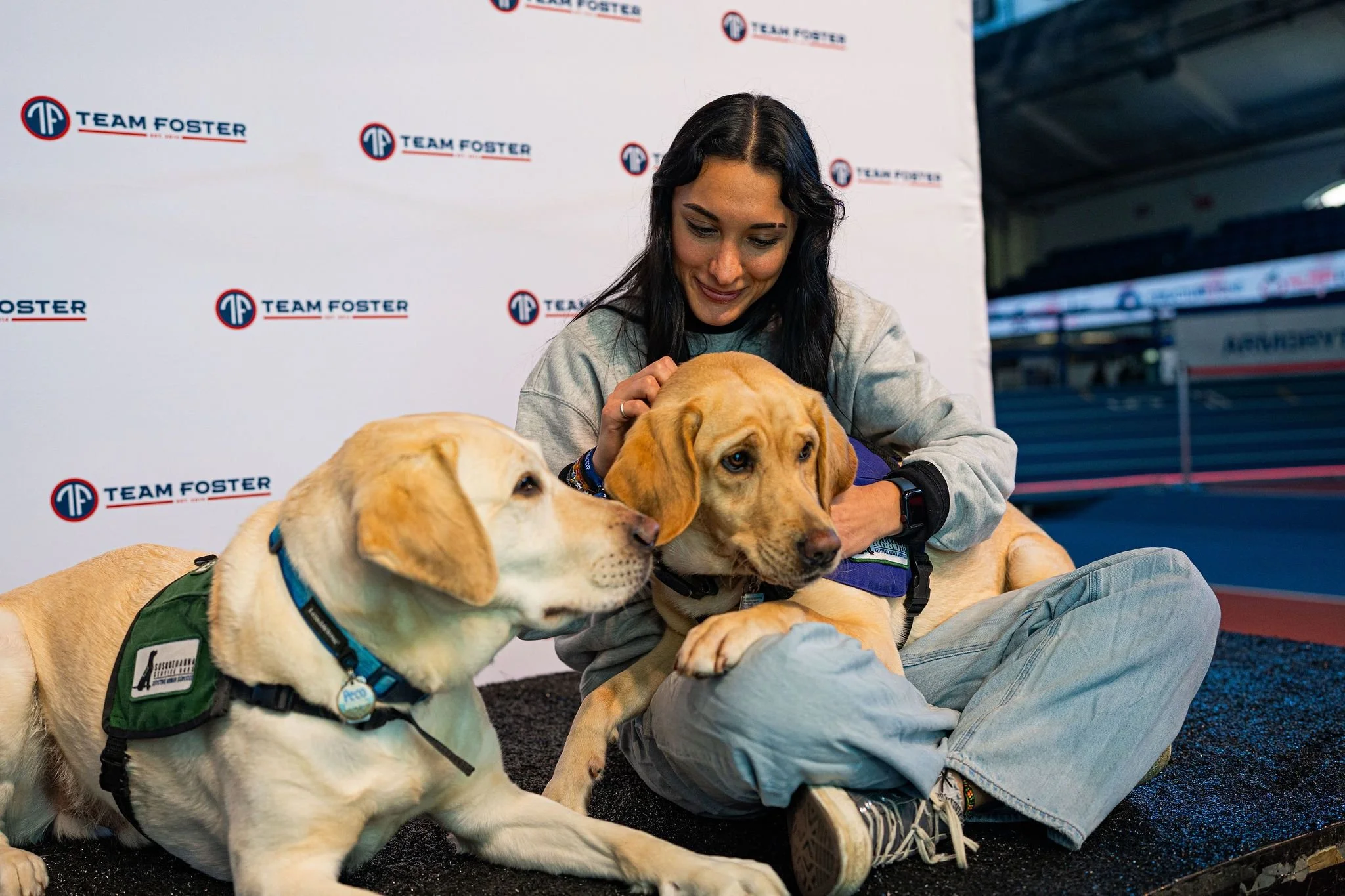 A woman sitting on the ground at an indoor event, surrounded by service dogs wearing harnesses, with a backdrop featuring the Team Foster logo.