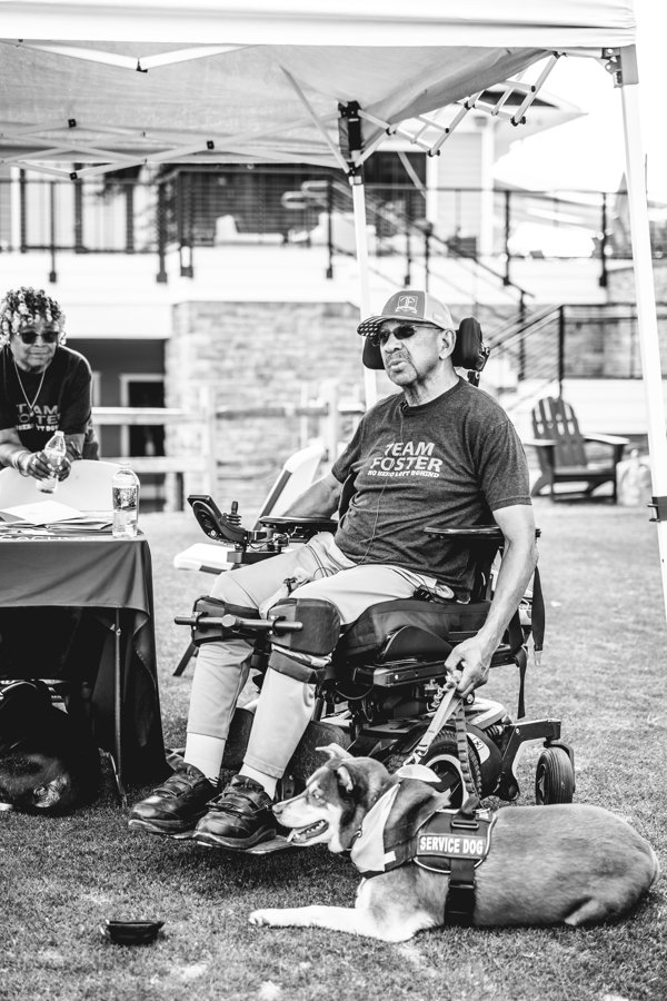 A man in a motorized wheelchair wearing sunglasses and a T-shirt that says "TEAM FOSTER" with a service dog lying on the grass beside him. The man is outdoors under a canopy, and there is a woman in the background at a table.