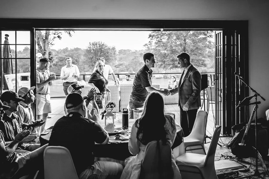 A group of people at a celebration, with two men shaking hands at an open doorway, some seated at tables, and others standing outside overlooking a scenic landscape.