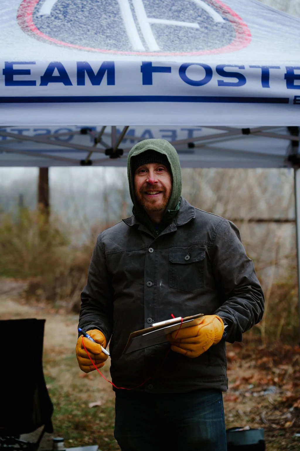 A man wearing a black jacket, green knit hat, and orange gloves standing outdoors under a tent with a sign that reads 'TEAM TOSTE'. He is smiling, holding a clipboard and a pen, and appears to be participating in an outdoor event in a wooded area.