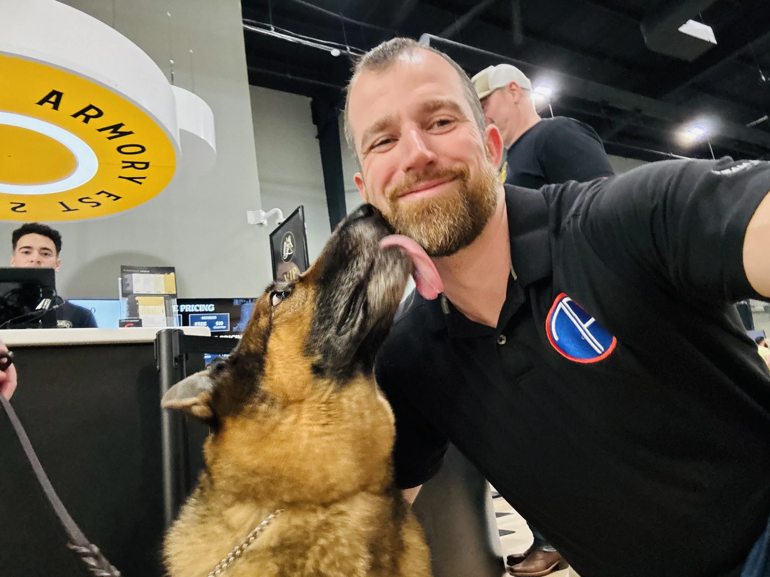 A man with a beard and a smile is hugging a dog. The dog is looking up at him with its tongue out. In the background, there is a sign that says 'ARMORY' and a man wearing a cap.