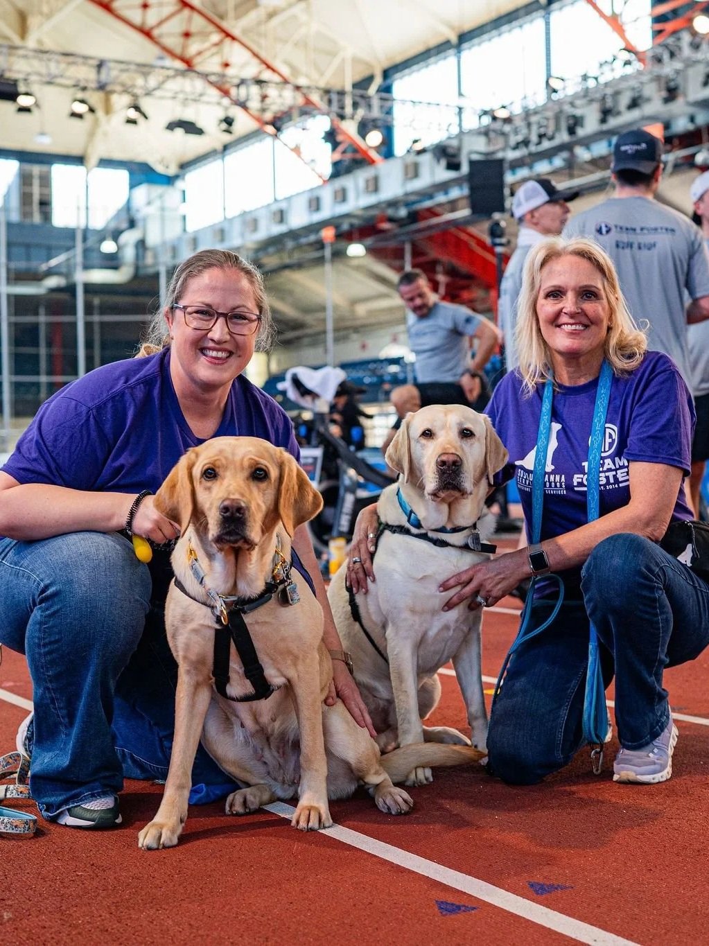Two smiling women kneel on an indoor track with two Labrador retrievers, surrounded by people and sports equipment in a large, well-lit indoor sports arena.