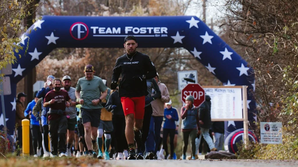 Group of people running through an outdoor event under a large blue archway that says 'Team Foster,' surrounded by trees.