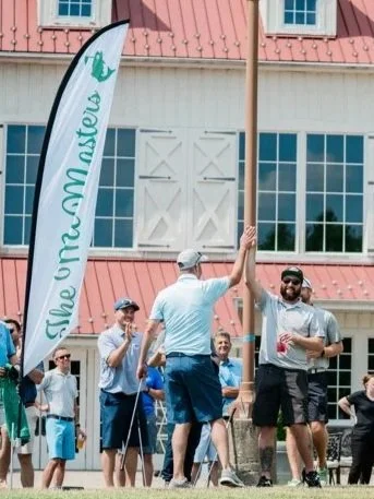 A golf event with people celebrating on a golf course, high-fiving near a flag that reads 'The Community Inistute' in front of a building with large windows.