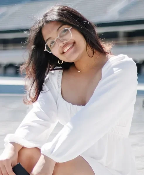 A young woman wearing glasses and a white dress smiling outdoors near boats.