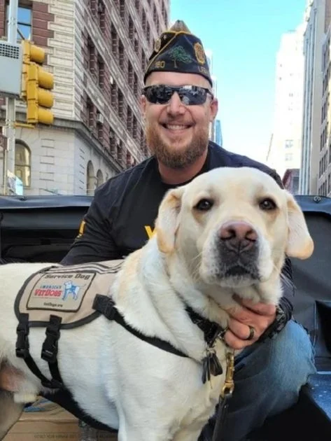 Man with a beard, sunglasses, and a World War II veteran cap smiling while holding a service dog in an urban setting.