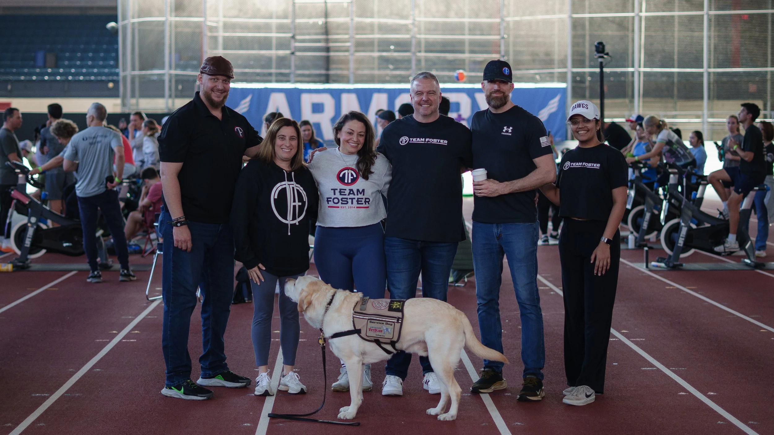 Group of six people, including a service dog, standing together in an indoor athletic facility, with exercise bikes and other people in the background.