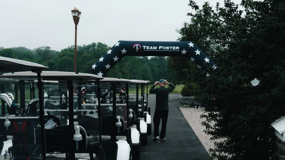 Golf carts lined up on a golf course pathway with an inflatable archway that reads 'Team Foster' and a person adjusting sunglasses in the background.