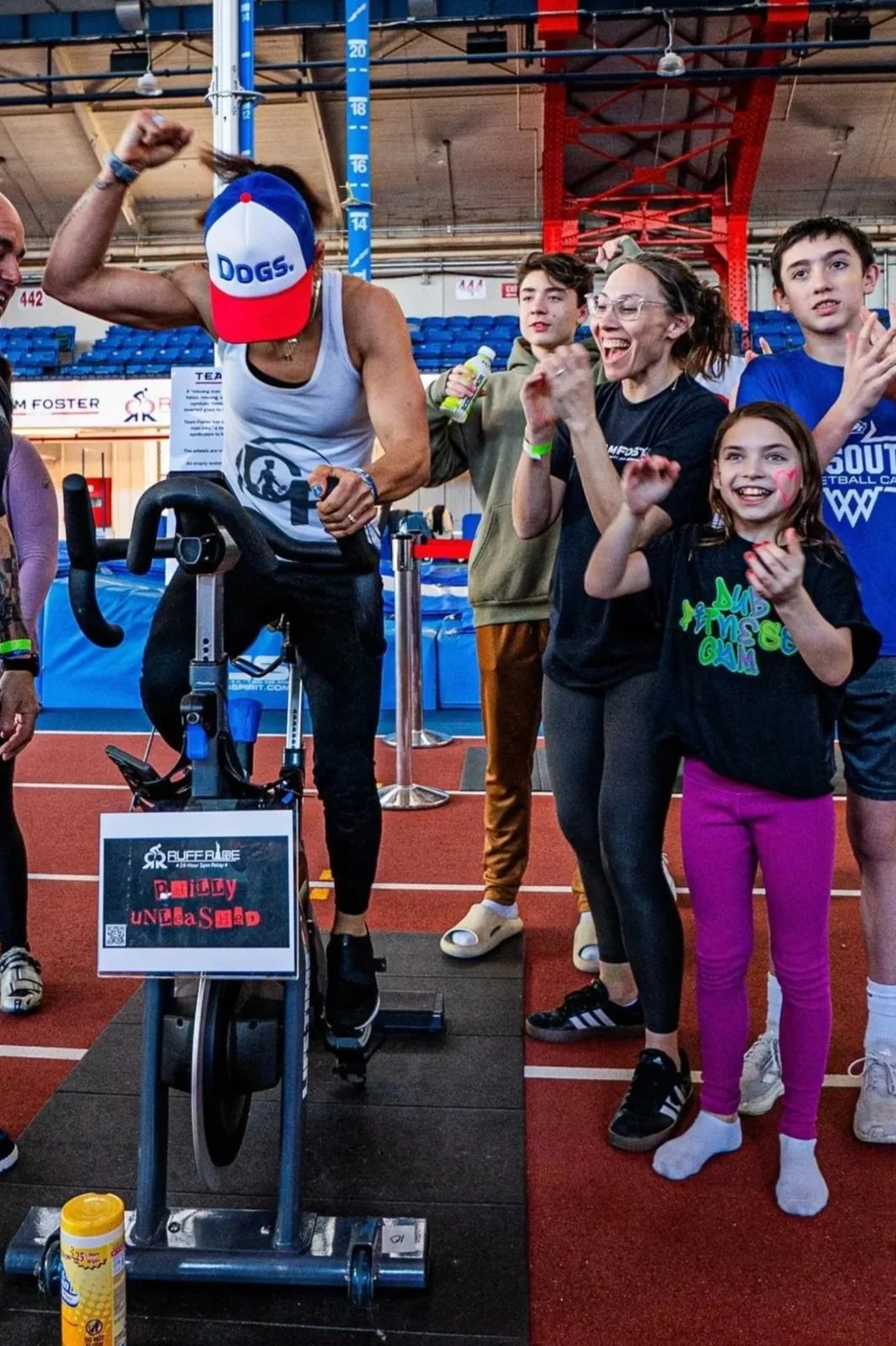 A woman in athletic attire flexing her arm on a stationary exercise bike, surrounded by smiling people in an indoor sports arena.