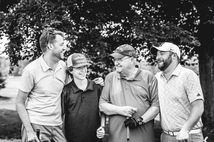 Four men standing outdoors, smiling, and talking to each other, with trees in the background.