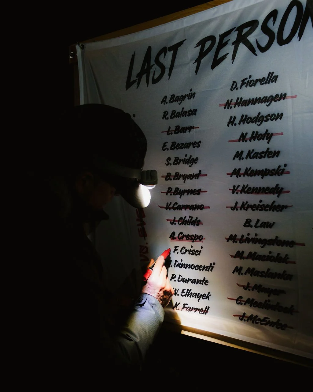Person illuminating a list of names on a whiteboard titled 'Last Person' with a headlamp