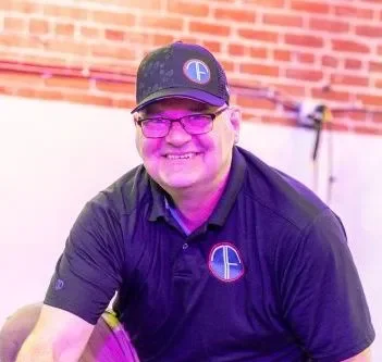 A smiling man wearing glasses, a black baseball cap with a logo, and a black polo shirt with a logo, sitting indoors with a brick wall in the background.