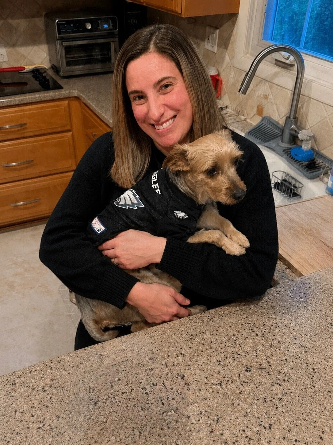 A woman smiling and holding a small dog in a kitchen. The woman is wearing a black long-sleeve shirt, and the dog is dressed in a black sports jersey. The kitchen has wooden cabinets, a countertop, a sink, a small oven, and a window with blue sky visible outside.