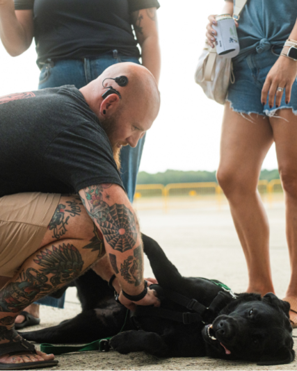 A man with tattoos and a bald head lying on the ground with a black dog, while people stand nearby holding drinks.
