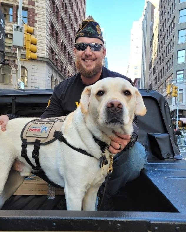 A smiling man with a beard, wearing sunglasses, a black jacket, and a navy cap, is holding a service dog in a city street setting. The service dog is a large, light-colored Labrador Retriever with a service vest.