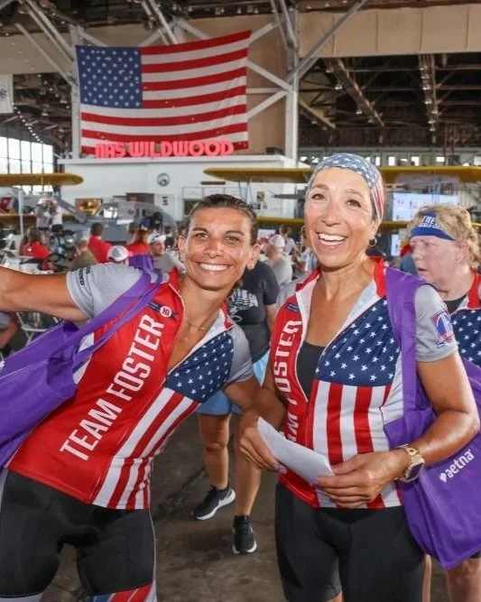 Two women wearing American flag-themed shirts and purple backpacks inside a large indoor venue, smiling and posing for the camera, with an American flag hanging in the background.
