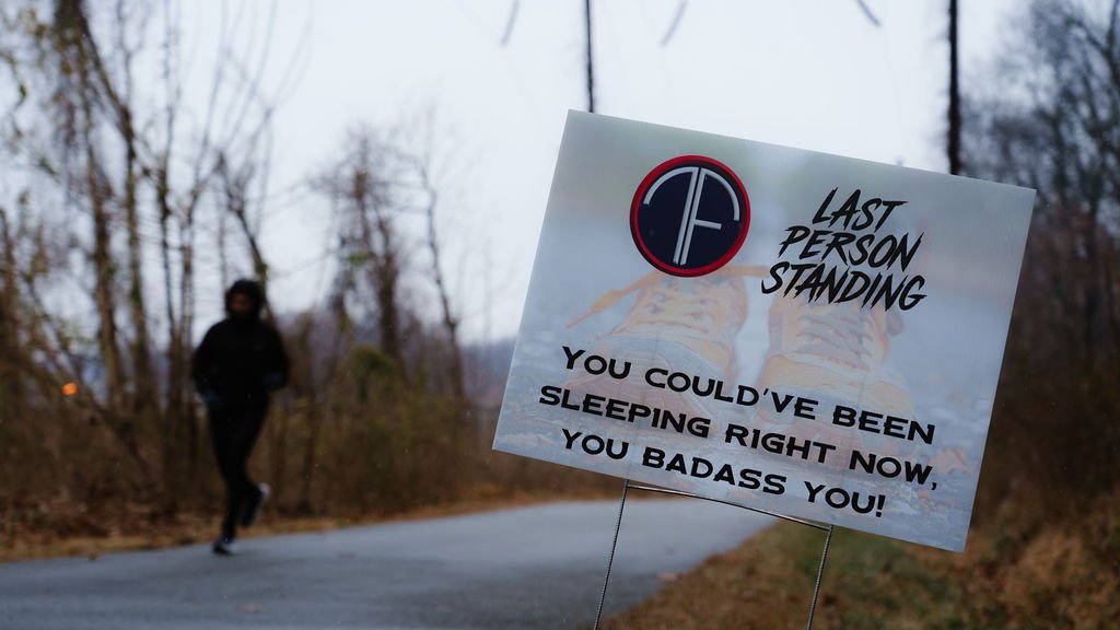 A person running on a rural road with a sign in the foreground that reads, "LAST PERSON STANDING. YOU COULD'VE BEEN SLEEPING RIGHT NOW, YOU BADASS YOU!" The background shows leafless trees and an overcast sky.