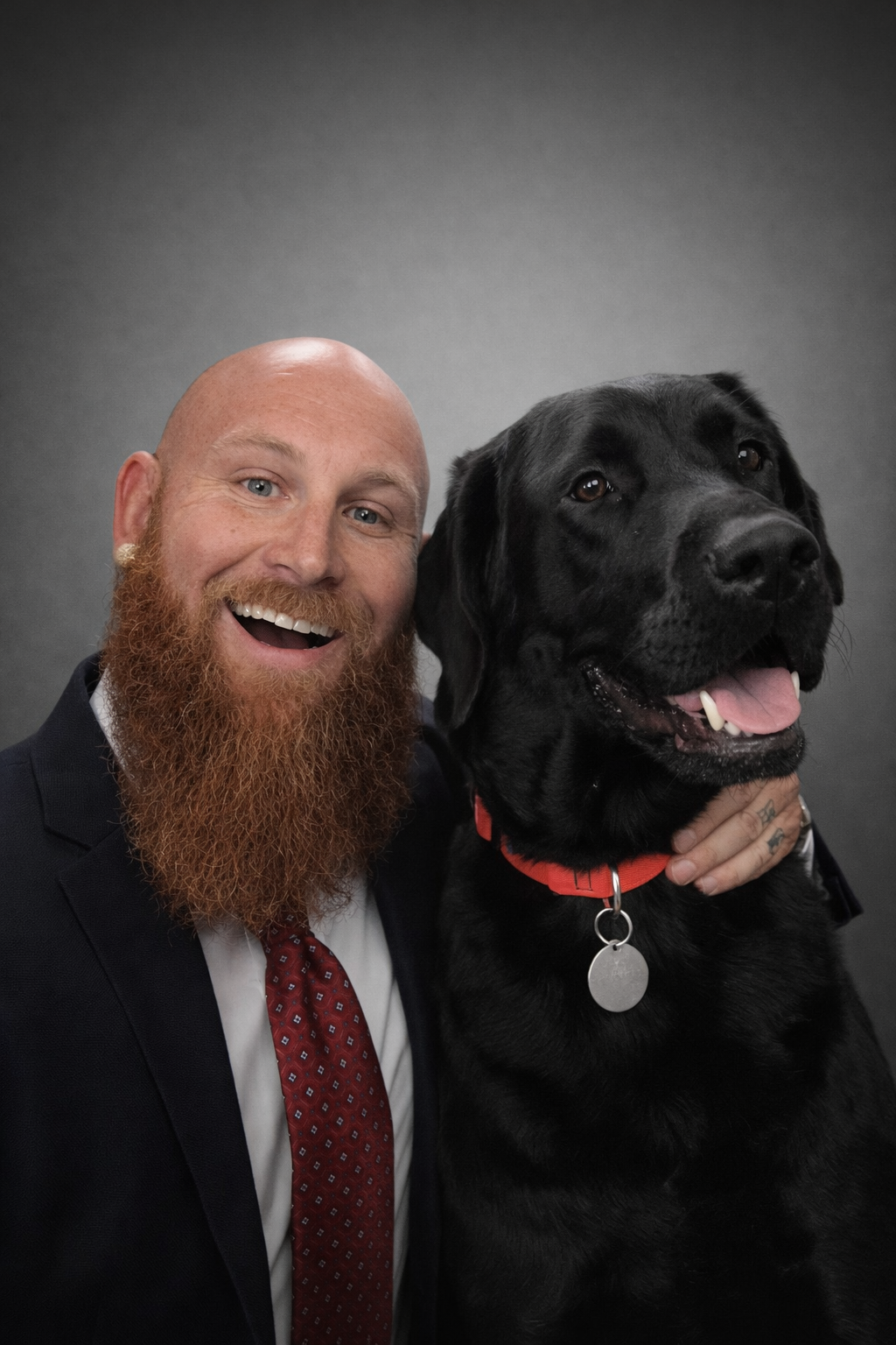 A smiling man with a long red beard and a bald head, wearing a suit and red patterned tie, posing with a black Labrador Retriever dog against a dark background.