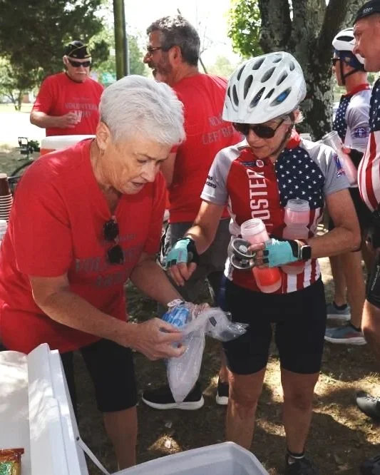 Group of people outdoors, some wearing red T-shirts and cycling gear, including a woman with a helmet, sunglasses, and gloves, holding a water bottle, in a park with trees in the background.